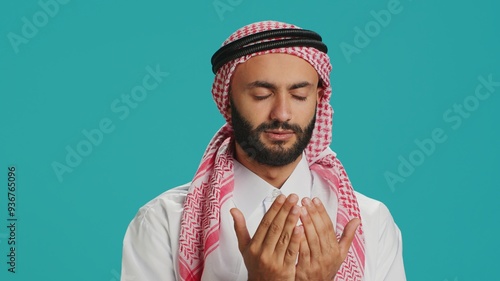 Muslim person praying to allah on camera, having belief in religious ritual over blue background. Middle eastern guy worshipping God with meditation, islamic cultural identity. photo