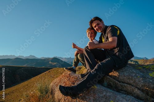 Father and son sitting on huge stone in the mountains at sunset light and showing thumbs up photo