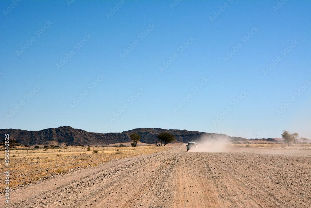 A car drives towards the hills on a dirt road in a desert area in perspective under a clear blue sky A car drives towards the hills on a dirt road in a desert area in perspective under a clear blue sky