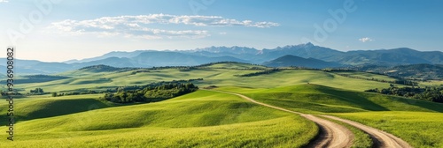 A scenic landscape view of rolling green hills and mountains under a blue summer sky, with a winding dirt road leading through the Italian countryside