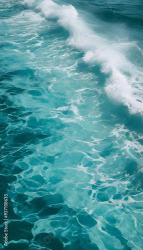 Aerial view of turquoise ocean waves crashing gently, creating white foam. The water is clear and vibrant, showcasing various shades of blue and green.