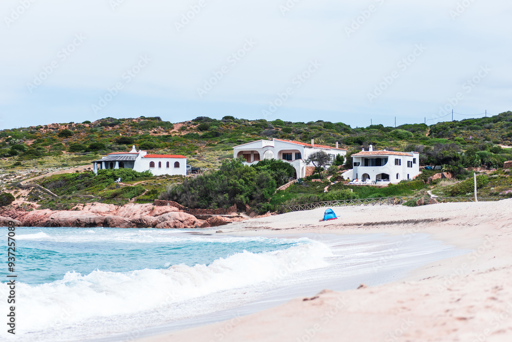 Fototapeta premium Overlooking the bay from Isola Rossa beach, the turquoise waters gently lap against the pink rocky coastline, where a few Mediterranean villas are nestled among the lush green hills.