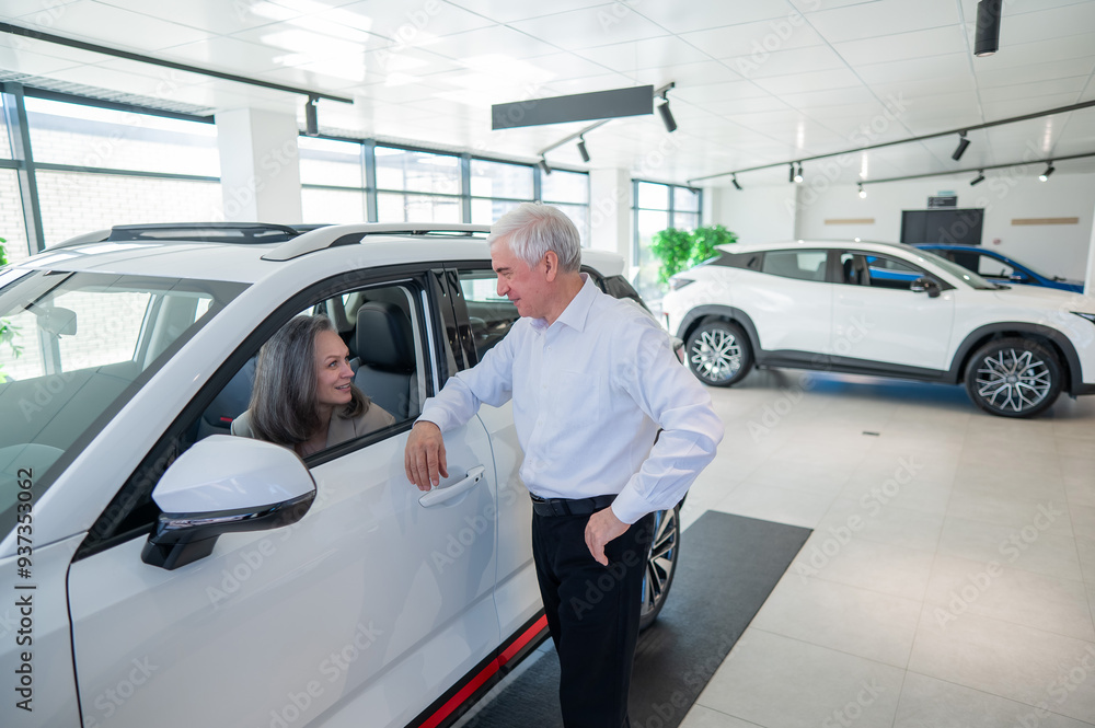An elderly Caucasian woman is sitting in a new car. Her husband is standing nearby. An elderly Caucasian woman is sitting in a new car. Her husband is standing nearby.