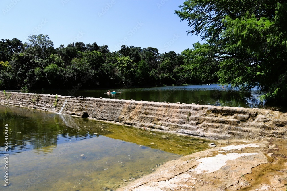 Serene river in the Blanco State Park with a small stone dam, surrounded by lush green trees Serene river in the Blanco State Park with a small stone dam, surrounded by lush green trees