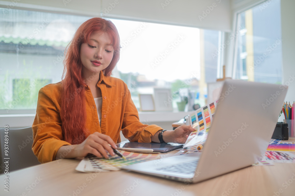 Young asian woman graphic designer with red hair is focused on her work, selecting colors for her project at desk. Young asian woman graphic designer with red hair is focused on her work, selecting colors for her project at desk.
