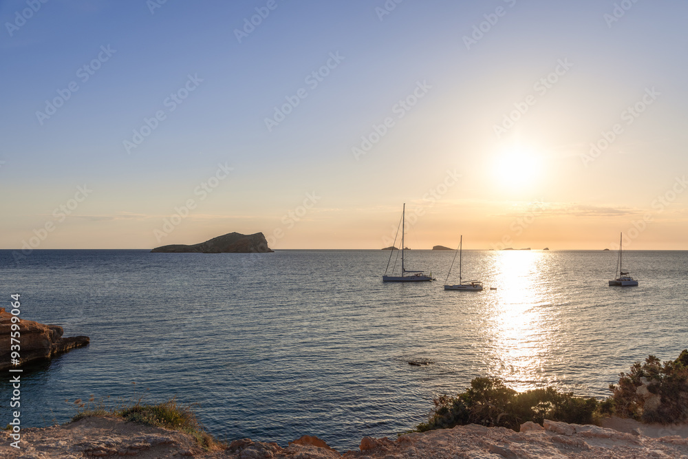 Serene sunset at Cala Conta beach in Ibiza with sailboats gently floating on the calm waters. The sun casts a golden glow over the horizon, illuminating the distant islands Serene sunset at Cala Conta beach in Ibiza with sailboats gently floating on the calm waters. The sun casts a golden glow over the horizon, illuminating the distant islands