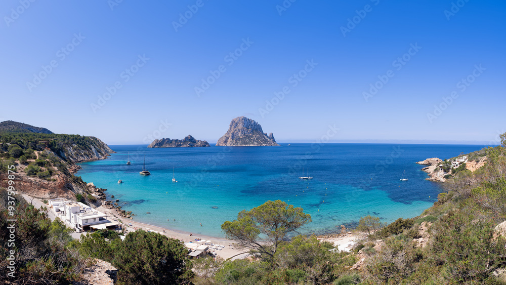 Expansive panorama of Cala d'Hort beach, Ibiza, showcasing Es Vedra island, tranquil turquoise waters, sailboats, and surrounding cliffs with vegetation, all beneath a bright sky Expansive panorama of Cala d'Hort beach, Ibiza, showcasing Es Vedra island, tranquil turquoise waters, sailboats, and surrounding cliffs with vegetation, all beneath a bright sky