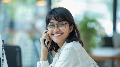 With a warm smile, a confident Indian woman talks on the phone at her stylish desk, surrounded by natural light and greenery in a contemporary office setting