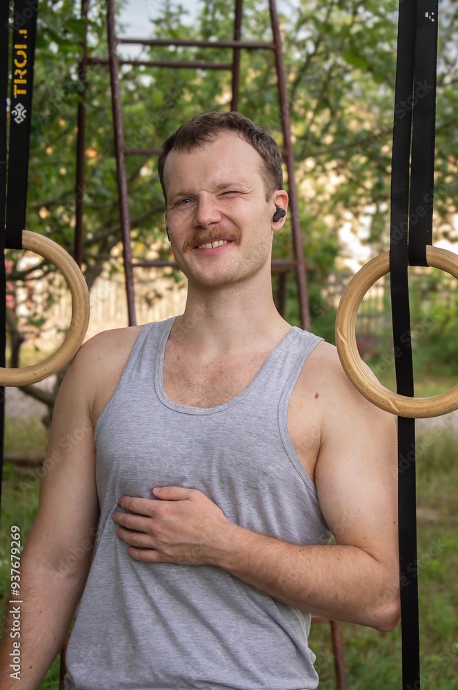 Obraz premium Winking man with a mustache standing between wooden gymnastic rings, wearing a gray tank top and wireless earbuds in an outdoor setting with trees.