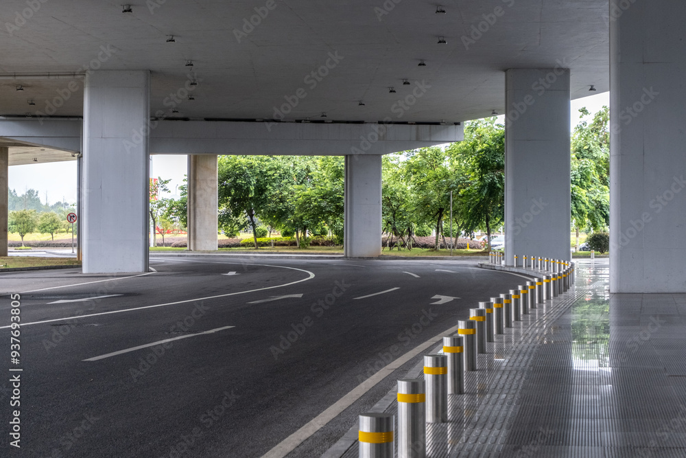 A curved road under a concrete overpass with thick pillars and a high ceiling, featuring a series of silver bollards along the side, separating from a pedestrian walkway in a modern urban setting A curved road under a concrete overpass with thick pillars and a high ceiling, featuring a series of silver bollards along the side, separating from a pedestrian walkway in a modern urban setting