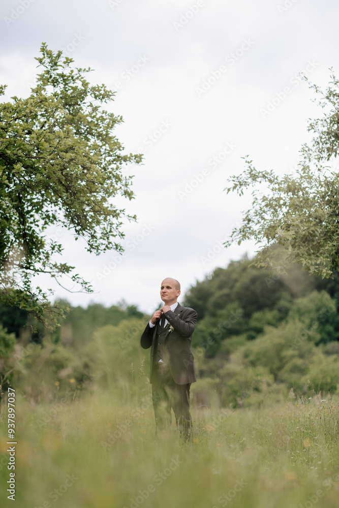 A man in a suit stands in a field of grass. The sky is cloudy and the man is looking up