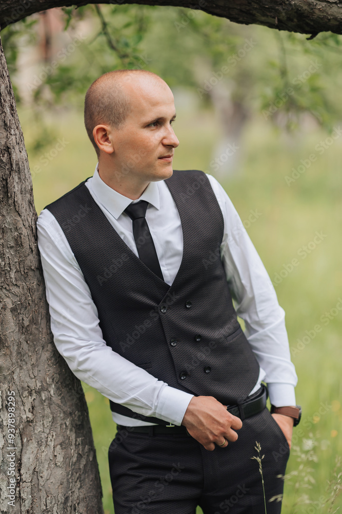 A man in a black vest and white shirt stands next to a tree. He is wearing a black tie and has his hands in his pockets. Concept of formality and elegance A man in a black vest and white shirt stands next to a tree. He is wearing a black tie and has his hands in his pockets. Concept of formality and elegance