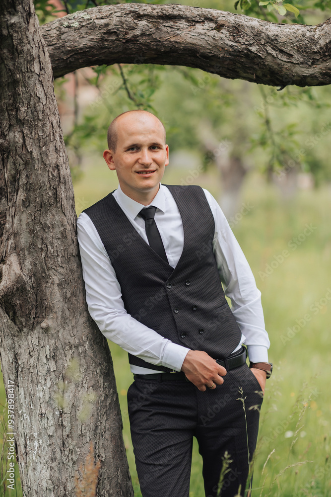 A man in a black vest and tie stands next to a tree. He is smiling and looking at the camera A man in a black vest and tie stands next to a tree. He is smiling and looking at the camera