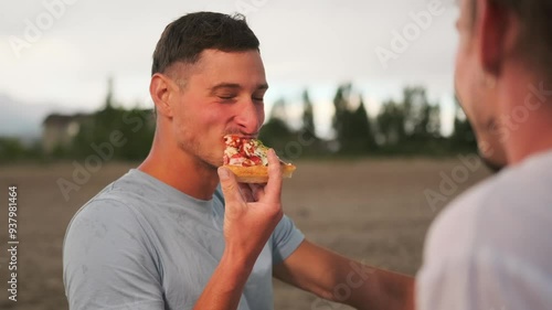 Hungry guy laughing eating pizza and looking at friend on nature during vacation. Slow motion, close up. photo