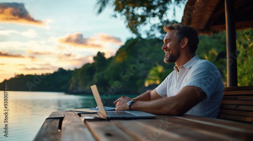 Fototapeta Naklejka Na Ścianę i Meble -  Smiling man working remotely with a laptop, seated by a lake during sunset, enjoying the tranquil outdoor environment.