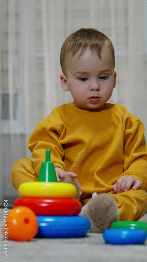 Adorable little baby in orange suit sits surrounded by toys. Cute child looks aside and pulls his sock from a foot. Vertical video