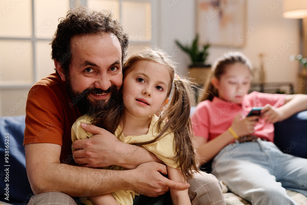 Father and daughter sharing affectionate moment on couch while another child using smartphone in background creating warm family atmosphere in cozy living room Father and daughter sharing affectionate moment on couch while another child using smartphone in background creating warm family atmosphere in cozy living room