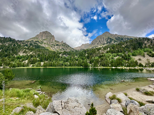 Lake Ratera is nestled among lush, green mountains in the Catalan Pyrenees. The calm waters reflect the surrounding landscape under a partly cloudy sky, offering a tranquil scene. photo