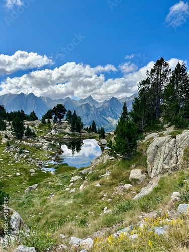 A small, clear lake near Amitges Refuge reflects the surrounding mountain peaks and a vibrant blue sky. The rocky landscape is dotted with trees, capturing the rugged beauty of the Catalan Pyrenees. photo