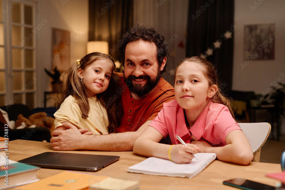 Smiling man sitting with his two daughters at table, helping them with homework. Younger girl leaning on father's shoulder while elder girl writing on notebook Smiling man sitting with his two daughters at table, helping them with homework. Younger girl leaning on father's shoulder while elder girl writing on notebook