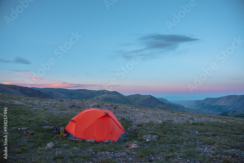 Orange tent on grassy stony pass with view to alpine valley and mountain range silhouette far away under clouds in sunset color sky. Red tent in high mountains under lenticular clouds in sunset sky.