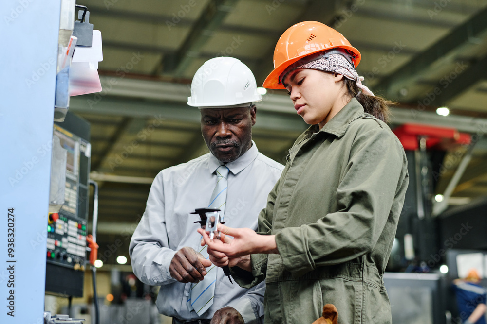 Two factory workers, one senior man and one young woman, discussing repair of machinery in an industrial setting, both wearing safety helmets and focused on task Two factory workers, one senior man and one young woman, discussing repair of machinery in an industrial setting, both wearing safety helmets and focused on task