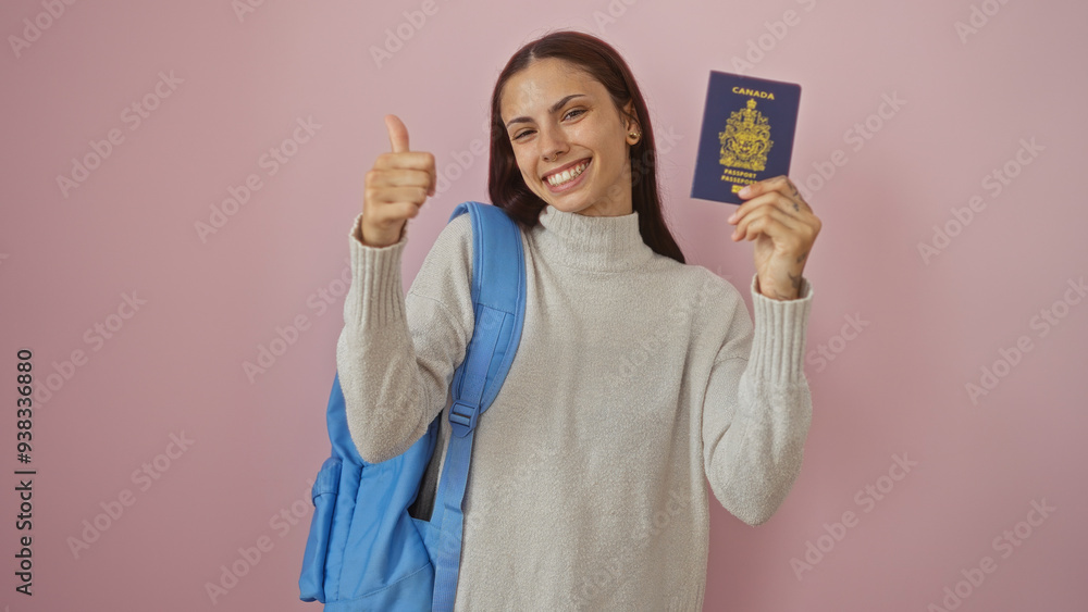 Young woman with brunette hair smiling and holding a canadian passport, giving a thumbs-up gesture, over a pink background, wearing a gray sweater and a blue backpack Young woman with brunette hair smiling and holding a canadian passport, giving a thumbs-up gesture, over a pink background, wearing a gray sweater and a blue backpack