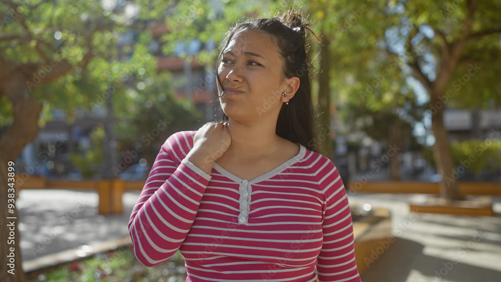 A young hispanic woman with a pained expression holds her neck in a leafy park setting, depicting discomfort or injury. A young hispanic woman with a pained expression holds her neck in a leafy park setting, depicting discomfort or injury.