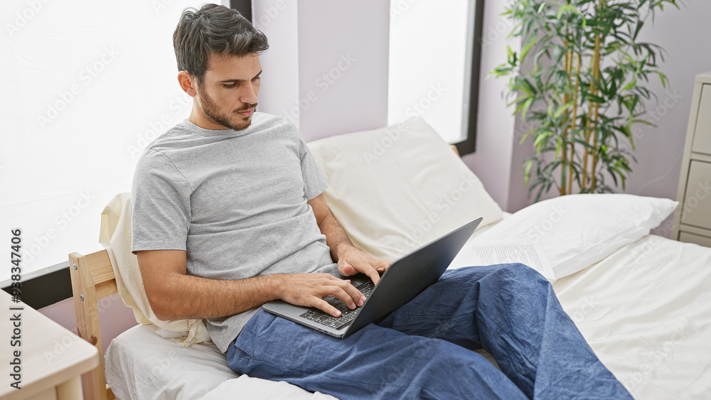 A young hispanic man with a beard works on his laptop while sitting on a bed in a well-lit bedroom. A young hispanic man with a beard works on his laptop while sitting on a bed in a well-lit bedroom.