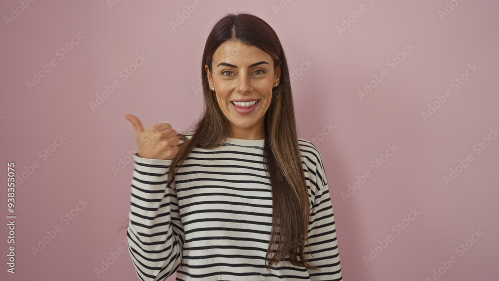 Smiling young hispanic woman in a striped shirt pointing with thumb against a pink isolated background Smiling young hispanic woman in a striped shirt pointing with thumb against a pink isolated background
