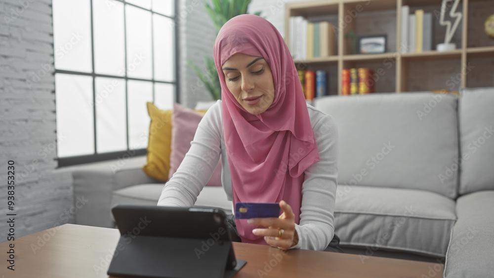 A focused woman in a hijab uses a credit card for an online transaction in a modern living room. A focused woman in a hijab uses a credit card for an online transaction in a modern living room.