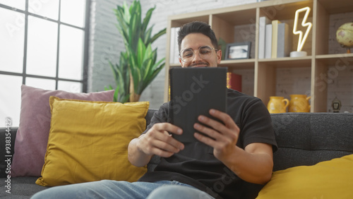 Handsome hispanic man with beard using tablet comfortably in a cozy, modern living room.