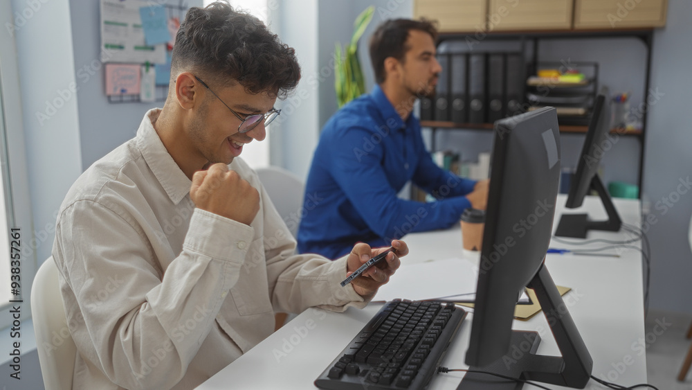 Men working in an office, one smiling at his phone while the other focuses on his computer, indicating a mix of productivity and a possible business success or achievement.