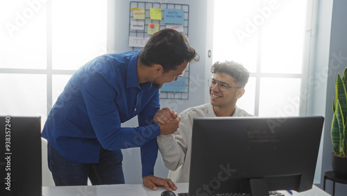 Hispanic male workers smiling and shaking hands in a modern office interior; one wearing glasses and casual attire seated, the other standing in a blue shirt near computers and charts