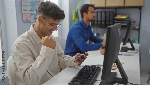 Wallpaper Mural Men working in an office, one smiling at his phone while the other focuses on his computer, indicating a mix of productivity and a possible business success or achievement. Torontodigital.ca