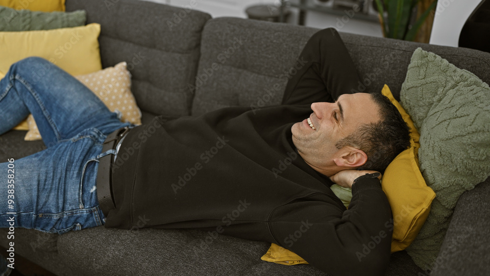 A relaxed hispanic man smiling while lying on a grey sofa with colorful pillows in a cozy living room setting. A relaxed hispanic man smiling while lying on a grey sofa with colorful pillows in a cozy living room setting.