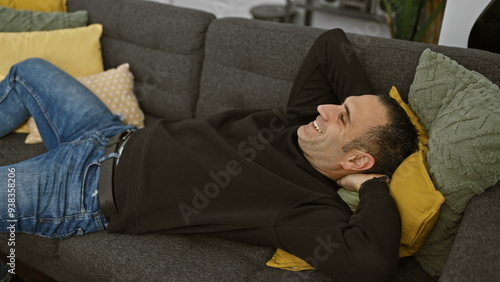 Wallpaper Mural A relaxed hispanic man smiling while lying on a grey sofa with colorful pillows in a cozy living room setting. Torontodigital.ca