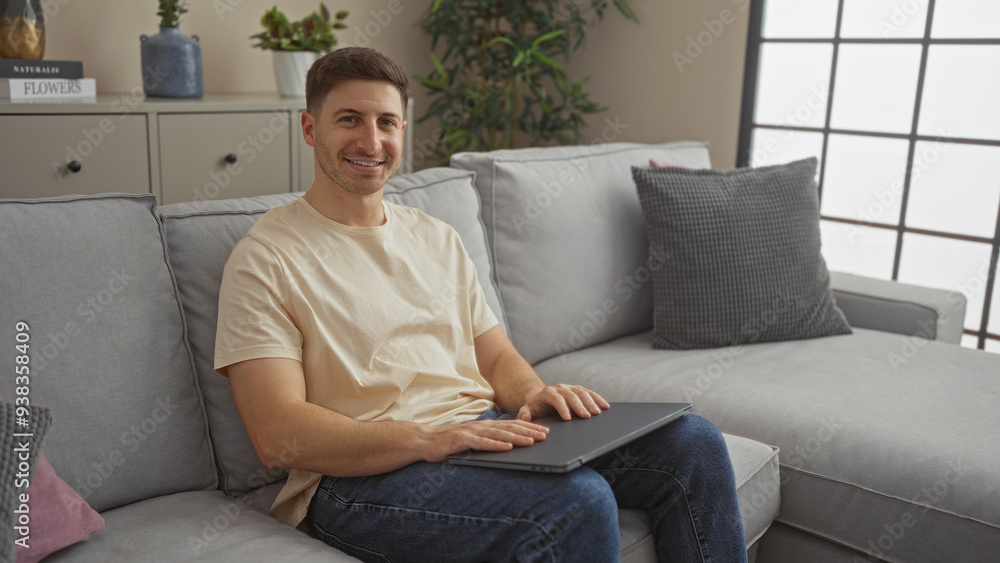 A handsome young man smiling indoors while sitting on a comfortable sofa in a modern living room apartment. A handsome young man smiling indoors while sitting on a comfortable sofa in a modern living room apartment.
