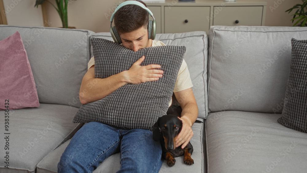 A young hispanic man with headphones holds a pillow on a grey couch in his living room, next to a small black dachshund dog, creating a cozy indoor atmosphere. A young hispanic man with headphones holds a pillow on a grey couch in his living room, next to a small black dachshund dog, creating a cozy indoor atmosphere.