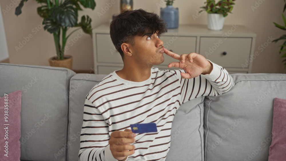 Young hispanic man in a striped shirt sitting on a couch in a cozy living room, holding a blue credit card, thoughtfully touching his lips, surrounded by indoor plants and soft furnishings.