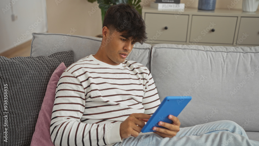 Handsome young hispanic man sitting in a living room, using a tablet and wearing a striped sweater, looks focused and relaxed, surrounded by a cozy, modern interior. Handsome young hispanic man sitting in a living room, using a tablet and wearing a striped sweater, looks focused and relaxed, surrounded by a cozy, modern interior.