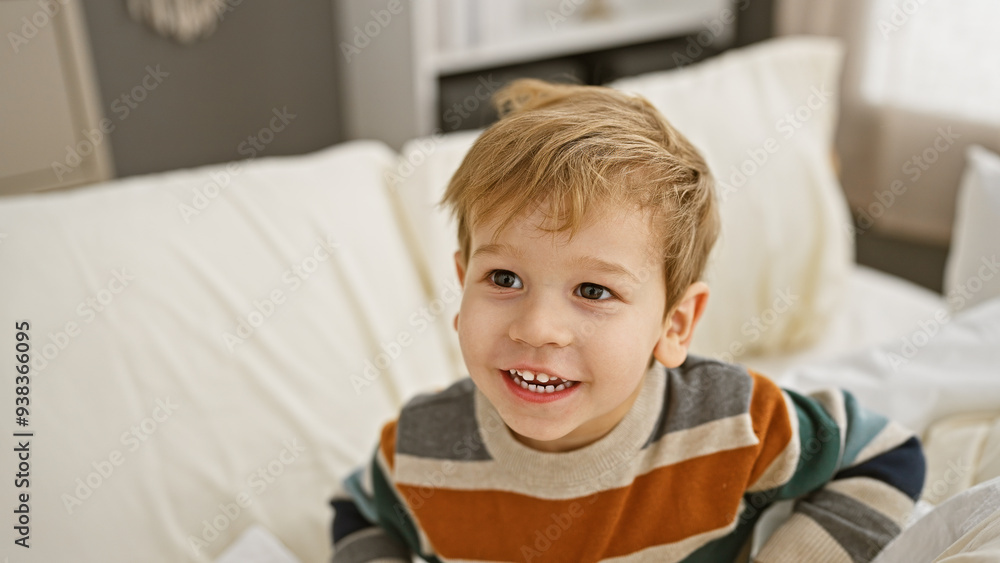 Smiling blond toddler boy sitting in a bright bedroom on a white bed, exuding childhood joy and innocence indoors. Smiling blond toddler boy sitting in a bright bedroom on a white bed, exuding childhood joy and innocence indoors.