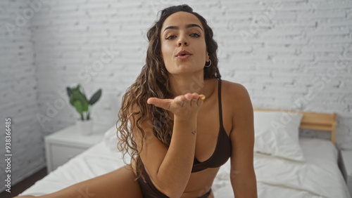 Tableau sur toile Young woman blowing a kiss while sitting on a bed in a cozy bedroom with white b