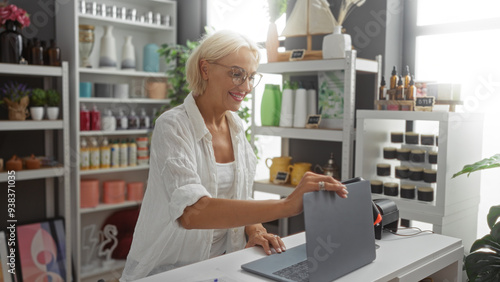 Woman smiling while working with laptop in home decor store, surrounded by various colorful decorations and plants