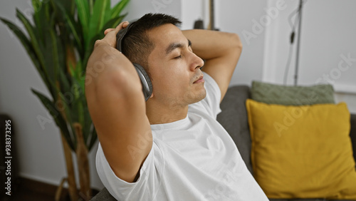 Relaxed hispanic young man enjoying music with headphones in a cozy living room interior.