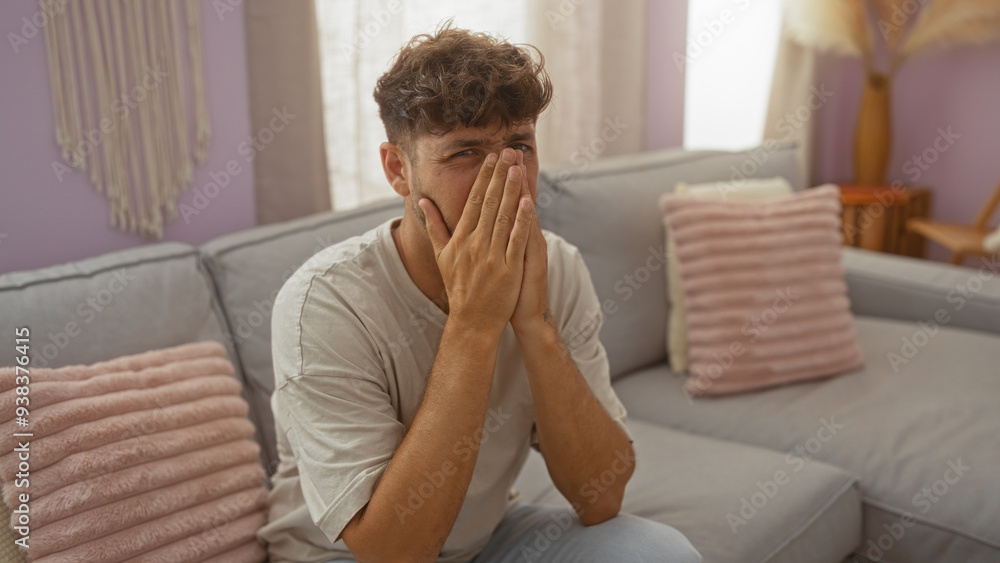 Young man sitting in a living room with his face covered, looking emotional and distressed in a modern, comfortable apartment setting. Young man sitting in a living room with his face covered, looking emotional and distressed in a modern, comfortable apartment setting.
