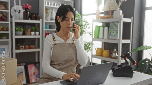Woman working in home decor store talking on phone using laptop surrounded by shelves filled with various home decoration items