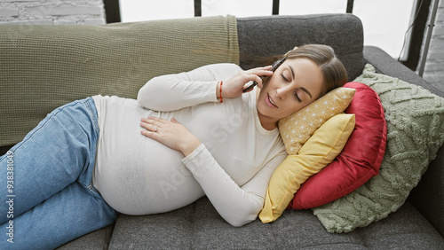 A young, pregnant woman relaxes at home on a couch, smiling as she talks on the phone, surrounded by colorful pillows.