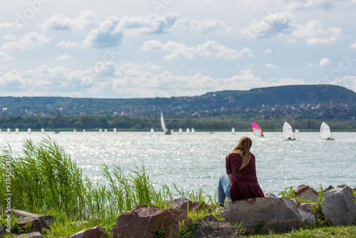 Young girl sitting on rocks watching ships and surfers sailing in the distance. Mountains and houses in the background. Spring or summer. Cloudy sky. Relaxation, reflection, dreams. Balaton, Hungary