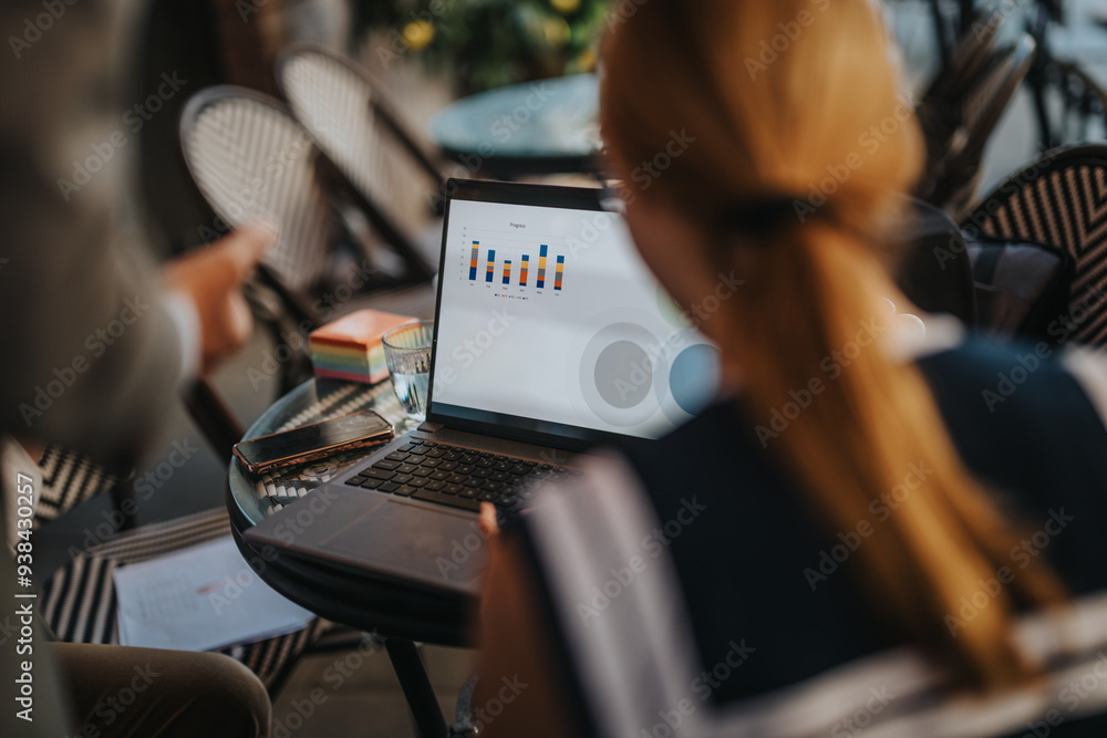 Two businesspeople review charts and data on a laptop in an outdoor cafe, highlighting teamwork, analysis, and business strategy. Two businesspeople review charts and data on a laptop in an outdoor cafe, highlighting teamwork, analysis, and business strategy.